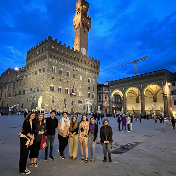 Students in front of the Palazzo Vecchio at night. The Palazzo and Loggia to the right are lit up, and the sky is a deep evening blue.