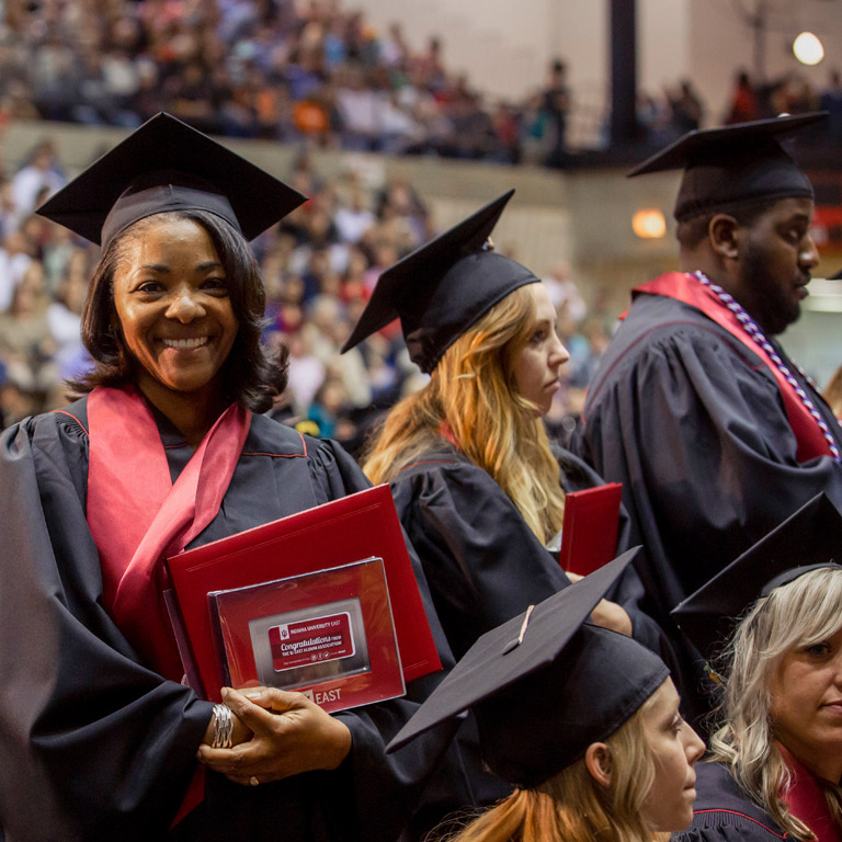 A middle-aged female IUE graduate holding graduation swag smiles from the crowd.
