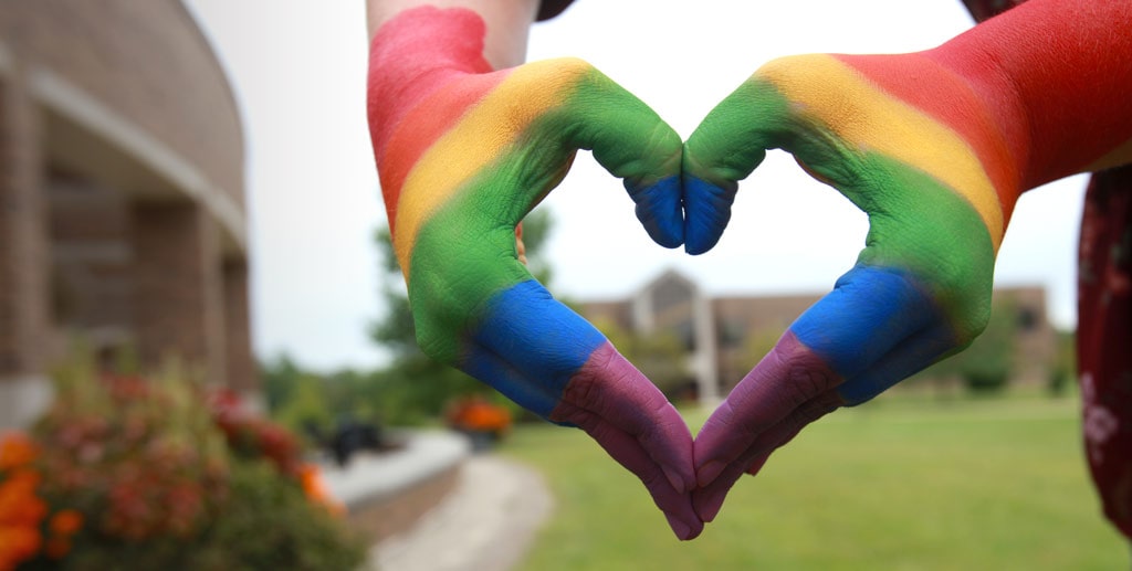 Hands, painted with the rainbow colors of the pride flag, making the shape of a heart with the IU East campus in the background.