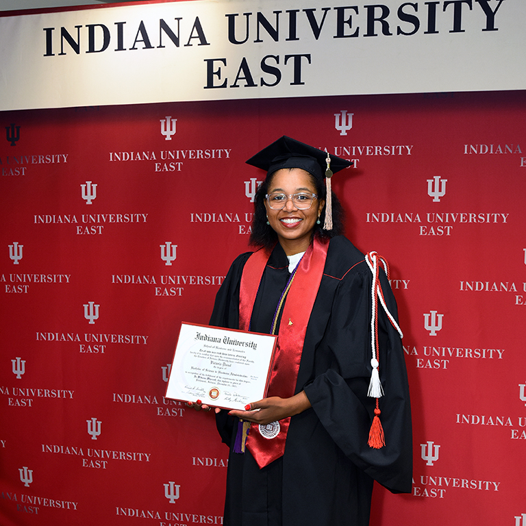 Vicky Duval stands with her diploma in front of the IU East media backdrop